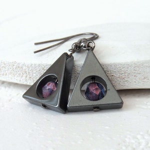 May include: A pair of geometric earrings. Each earring has a dark grey, triangular frame with a circular opening. Inside each opening is a faceted, purple bead. The earrings hang from dark grey ear wires.