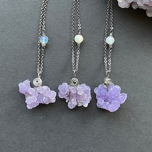 May include: Three necklaces with lavender-colored, clustered stone pendants. Each pendant is wire-wrapped and hangs from a silver chain. The necklaces are displayed on a dark gray surface, with green foliage in the background.