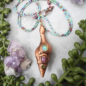 Peut inclure: Un collier avec un pendentif en forme de coquille de couleur cuivre. Le pendentif est orné de trois pierres précieuses : une verte, une rose et une bleu-verte. Le collier est composé de perles multicolores.