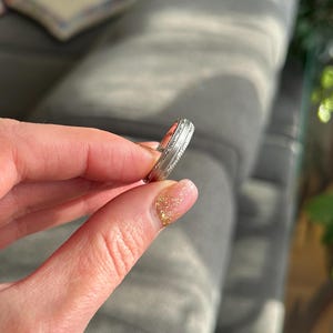 May include: A silver wedding band with a decorative design. The band is shown on a finger.