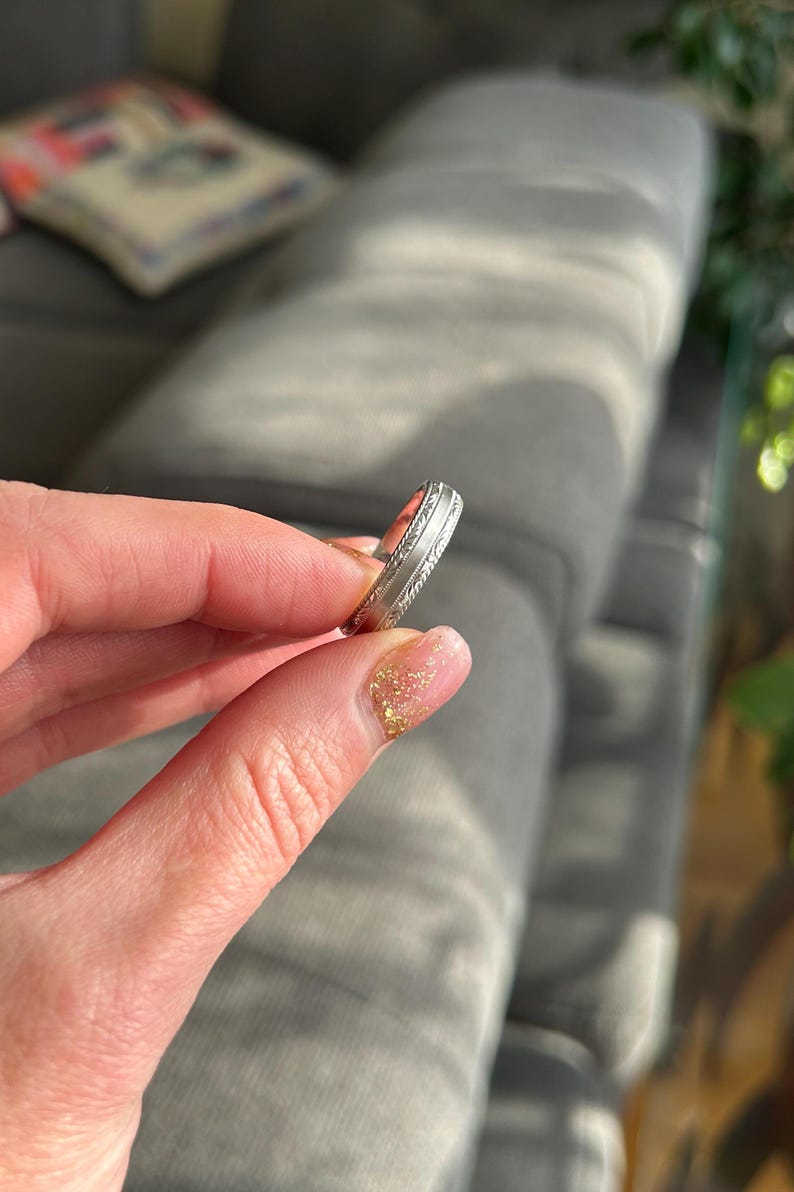 May include: A silver wedding band with a decorative design. The band is shown on a finger.