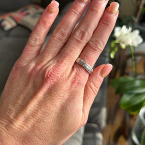 May include: A silver band ring with a textured design. The ring is worn on the ring finger of a person's hand.