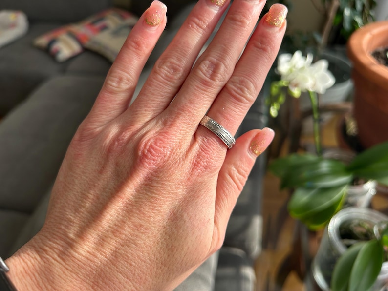 May include: A silver band ring with a textured design. The ring is worn on the ring finger of a person's hand.