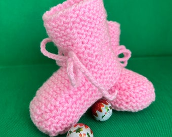 May include: Pink knitted baby booties with white laces. The booties are sitting on a green surface with three small, white and red Christmas ornaments.