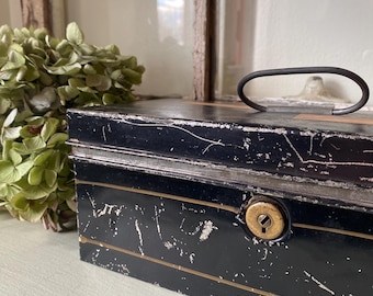 May include: A vintage black metal cash box with a handle and keyhole. The box has gold trim and shows signs of wear, with scratches and paint loss. A bunch of dried green flowers sits to the left of the box.