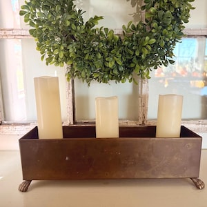 May include: A rectangular bronze-toned metal container with four small claw-style feet, holding three off-white pillar candles of varying heights. The container has a smooth, solid surface and is positioned in front of a green wreath.