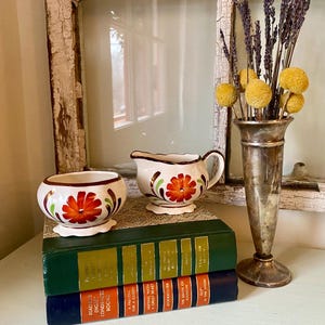 May include: A white ceramic creamer and sugar bowl set with orange floral designs and brown trim. The set sits atop a stack of vintage books. A silver vase holds dried flowers, and the background includes a window.