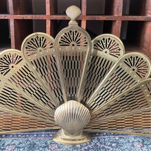 May include: A brass fireplace screen with a fan-shaped design. The screen features a central shell-shaped base and a series of radiating, perforated panels. The panels have a decorative pattern and are topped with a small shell-shaped finial.