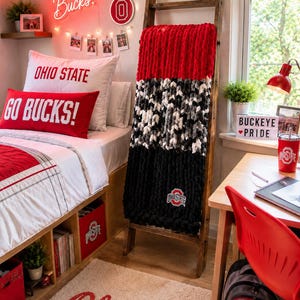 May include: A chunky knit throw blanket with horizontal stripes of red, a variegated mix of black, white, and gray, and solid black, featuring an Ohio State logo patch, draped over a wooden ladder in a room decorated with Ohio State themed items.