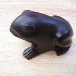 May include: A dark brown, carved wooden frog figurine. The frog has a smooth, polished surface and detailed features, including carved eyes and webbed feet. The figurine is set against a light wood grain background.