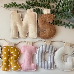 May include: A decorative banner spelling "MS BRIGGS" with fabric letters. The letters are in various colors and patterns, including white, yellow polka dots, pink, gray stripes, and brown. The banner is strung on twine.