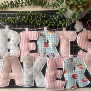 May include: A fabric banner spelling "Betsy's Bakery" in various patterns. The letters are in pink and blue, with star, stripe, and cupcake designs. The banner is strung on a pink and white twine, with a green plant in the background.
