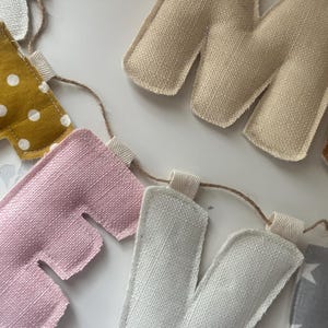 May include: A handmade fabric banner spelling out "HOME". The letters are in various colors, including pink, white, gold with white polka dots, and tan. The banner is strung on a natural twine.