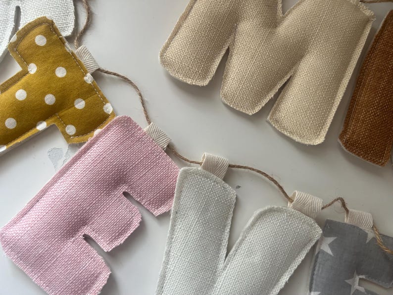 May include: A handmade fabric banner spelling out "HOME". The letters are in various colors, including pink, white, gold with white polka dots, and tan. The banner is strung on a natural twine.