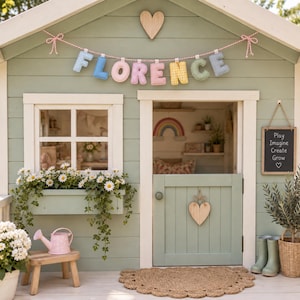May include: A sage green wooden playhouse featuring a fabric banner spelling FLORENCE in pastel colors, a window box with white daisies, a Dutch door with a wooden heart, and a small chalkboard sign with the text Play Imagine Create Grow.