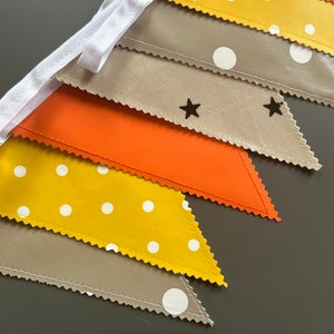May include: A decorative bunting banner featuring triangular flags in various colours. The flags are yellow with white spots, orange, beige with black stars, and beige with white spots. The banner is strung on a white ribbon.