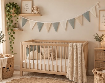 May include: A light wood crib with vertical slats, featuring a white mattress, a cream-colored knit throw blanket, and decorative pillows. Above the crib hangs a fabric bunting banner in alternating shades of sage green and cream.
