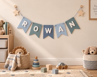 Op de afbeelding: Een decoratieve wandbanner met vijf blauwe en blauwgroene wimpels met witte houten letters die de naam ROWAN spellen. De banner hangt aan een zwart-wit koord dat is bevestigd aan twee lichtgekleurde houten wandhaken in de vorm van sterren.
