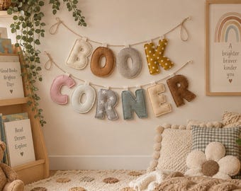 May include: A decorative wall banner featuring plush, fabric-covered letters that spell out the words BOOK CORNER. The letters are suspended on two separate twine strings and display a variety of colors and patterns, including solid tones, polka dots, and stars.