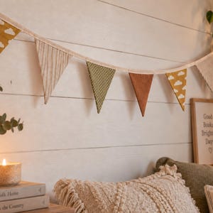 May include: A fabric bunting banner featuring a series of triangular flags in alternating patterns and colors, including mustard yellow with white clouds, beige stripes, olive green with white dots, and solid rust orange, all attached to a cream cord.