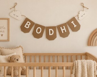 Op de afbeelding: Een decoratieve wandbanner met vijf bruine wimpels met witte letters die BODHI spellen, geflankeerd door twee witte wolkenuitsparingen met gesloten ogen. De banner is opgehangen aan beige touw, wat zorgt voor een horizontale weergave van de naam.