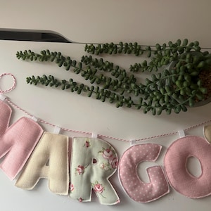 May include: A fabric banner spelling "MARGOT" in various colors and patterns. The letters are in pink, cream, and floral prints. The letters are strung on red and white twine. A small potted succulent plant is in the background, adding a touch of greenery.