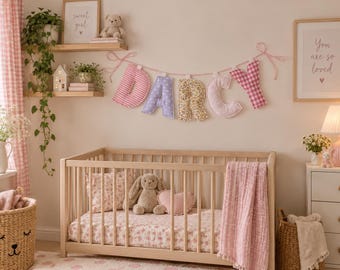 May include: A fabric banner spelling the name DARCY in individual stuffed letters, each featuring a unique pattern including pink stripes, purple stars, yellow floral print, solid light pink, and pink gingham, hung on a pink and white string.