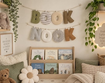 Puede incluir: Una decoración de pared con letras de tela que forman las palabras BOOK NOOK, colgadas de dos cuerdas. Las letras presentan diversos colores y estampados, incluyendo verde oliva, rayas grises, beige, marrón, gris y lunares blancos.