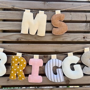 May include: A decorative banner spelling "MS BRIGGS" with fabric letters. The letters are in various colors and patterns, including cream, brown, yellow polka dots, pink, and gray stripes. The banner hangs on a wooden background.