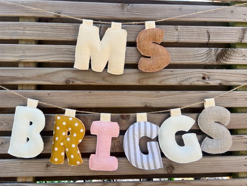 May include: A decorative banner spelling "MS BRIGGS" with fabric letters. The letters are in various colors and patterns, including cream, brown, yellow polka dots, pink, and gray stripes. The banner hangs on a wooden background.