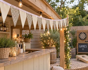 May include: A string of triangular bunting flags featuring a repeating white star pattern on a light beige fabric. The pennants are suspended from a white ribbon and hang across a wooden outdoor bar structure illuminated by warm string lights.