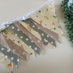 Puede incluir: Una bandera de banderines con temática de animales de safari. La bandera está hecha de tela beige, verde y blanca con una variedad de patrones, incluyendo lunares, estrellas e estampados de animales. La bandera está enhebrada con una cinta blanca.