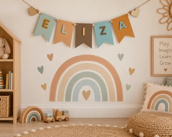 May include: A wall banner featuring the name ELIZA in cutout letters on multi-coloured pennants, suspended above a large rainbow wall decal with heart accents. The colour palette includes muted tones of sage green, terracotta, and brown.