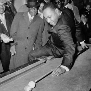 May include: Black and white photograph of a man in a suit playing pool. He is leaning over the pool table, aiming with a cue stick at the billiard balls. Several people are watching in the background.