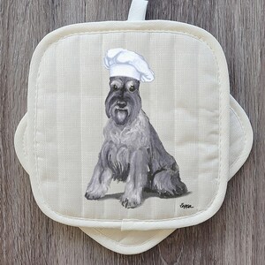 May include: Off-white, square pot holder with a painted Schnauzer dog wearing a chef's hat. The dog is gray and black, with a long beard and eyebrows. The pot holder has a striped pattern and a loop for hanging.