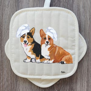 May include: Cream-colored, quilted oven mitt featuring a painted image of two corgi dogs wearing chef hats. One corgi is black, brown, and white, and the other is brown and white. The mitt has a loop for hanging.
