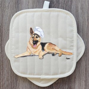 May include: Off-white, quilted pot holder with a German Shepherd wearing a chef's hat. The dog is painted in shades of tan, black, and white, with its tongue sticking out. The pot holder has a loop for hanging.
