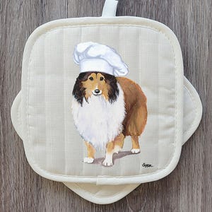 May include: Off-white, square pot holder with a painted image of a Collie dog wearing a chef's hat. The dog is brown, white, and black. The pot holder has a loop for hanging and a striped pattern.