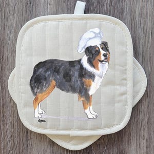 May include: Off-white, square pot holder with a watercolor painting of an Australian Shepherd wearing a chef's hat. The dog is black, brown, and white. The pot holder has a loop for hanging.
