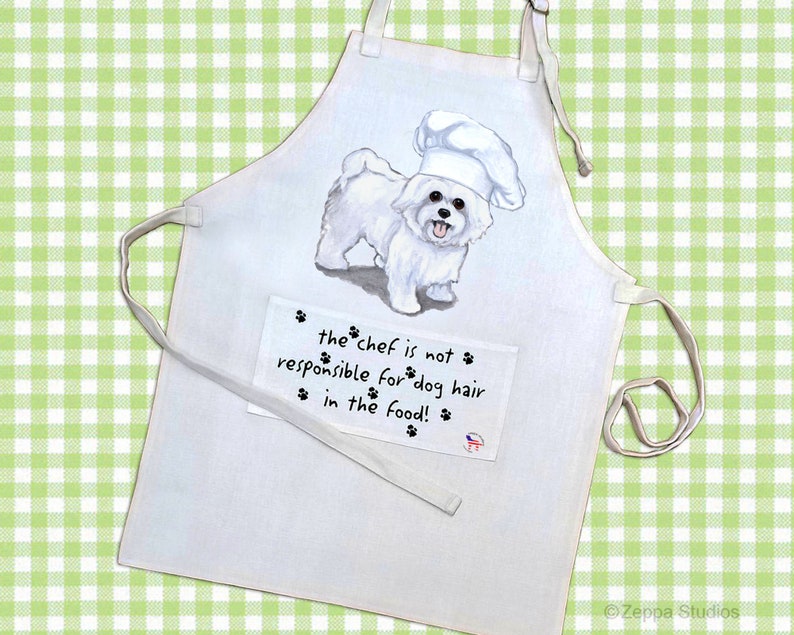 May include: White cotton apron with a white dog wearing a chef's hat. The apron has a pocket with the text "the chef is not responsible for dog hair in the food!"