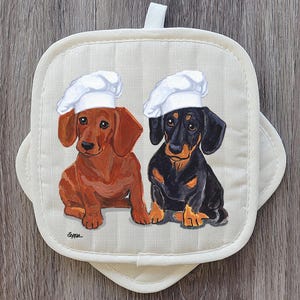 May include: Cream-colored oven mitt featuring a charming illustration of two dachshunds wearing chef hats. One dachshund is brown, the other is black and tan. The mitt has a loop for hanging.