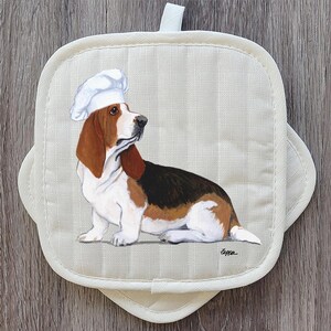 May include: Off-white square pot holder with a painted image of a Basset Hound wearing a chef's hat. The dog is brown, black, and white. The pot holder has a loop for hanging and a scalloped edge.