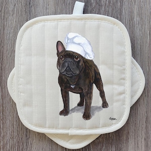 May include: Cream-colored, quilted pot holder with a French bulldog wearing a chef's hat. The dog is brown with a white patch on its chest. The pot holder has a loop for hanging.