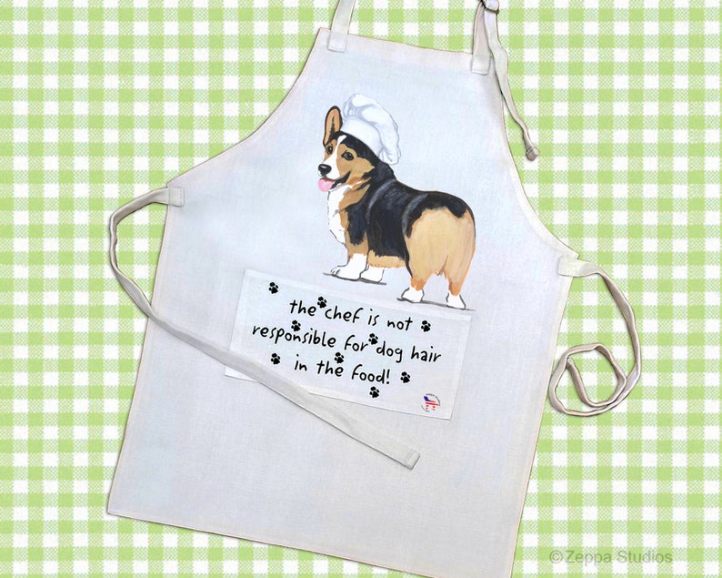 May include: White kitchen apron with a cartoon image of a corgi wearing a chef's hat. The text on the apron reads "the chef is not responsible for dog hair in the food!"
