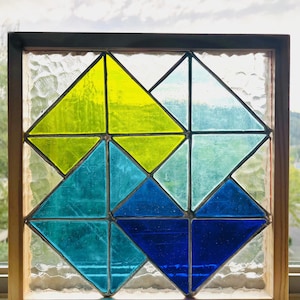 May include: Stained glass window with a geometric design. The window features a pattern of colorful triangles in shades of yellow, teal, and blue, set within a wooden frame. The glass has a textured appearance, and the window is positioned in front of a bright, natural background.