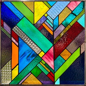 Könnte beinhalten: Ein farbenfrohes Buntglasfenster mit geometrischem Design. Das Kunstwerk zeigt eine Mosaik aus Farben, darunter Blau, Grün, Gelb, Rot und Rosa. Die Glasscheiben sind in einem Metallrahmen gefasst.