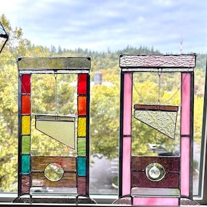 May include: Two stained glass guillotine decorations. One features a colorful design with red, yellow, and green hues. The other has a pink frame and a decorative glass blade. Both hang from chains.