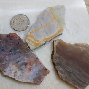 May include: Three pieces of polished agate, each with a unique pattern and color. The agate on the left is a mix of pink, white, and gray with black flecks. The agate in the center is white with yellow veins. The agate on the right is a light brown with white streaks.