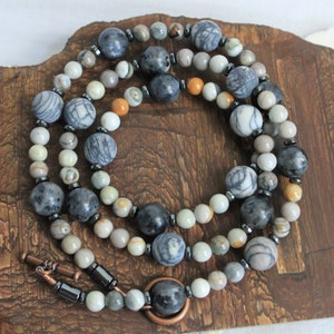 May include: A multi-strand necklace made with grey, white, and brown patterned beads. The necklace has a copper clasp and a copper ring.