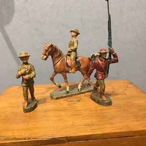 May include: Three wooden toy soldiers, one on horseback, one holding a flag, and one standing with a rifle. The soldiers are painted in brown, red, and green.